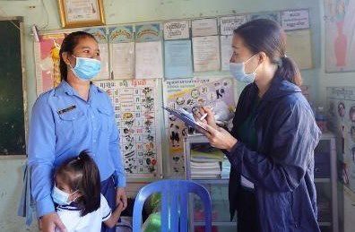 A preschool teacher in uniform speaks with a project staff member who takes notes in a classroom