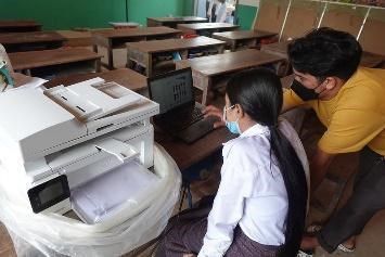 A teacher and a trainer set up a laptop beside a new printer in a classroom