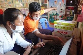 A trainer in an orange shirt shows a teacher how to use a laptop in a classroom