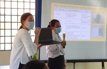 Two women hold a microphone and a laptop in front of a projected slide during the meeting
