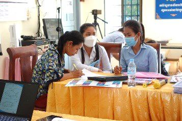 Two teachers write notes at a workshop table beside a laptop and printed game materials