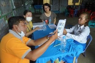 A staff member in an orange shirt shows a printed sheet to a teacher at a table during the assessment