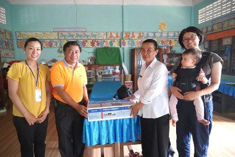Several people in yellow and orange shirts pose with a boxed laminator on a table in a classroom