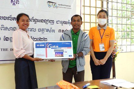 Three people hold a boxed laminator in front of a banner at a school