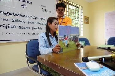 A teacher holds up a picture book to the room while a trainer in an orange shirt stands behind her