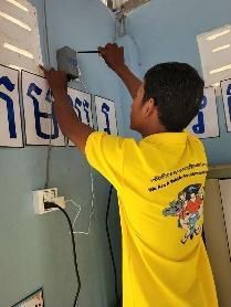 A staff member in a yellow shirt installs an AccessBox on a classroom wall near a power socket