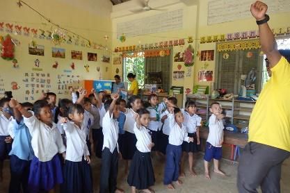 Young students in school uniform raise their arms and dance in their classroom as a staff member joins in
