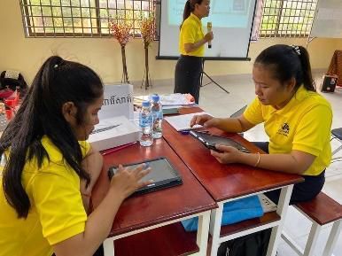 Teachers at a table use tablets while a trainer presents in the background
