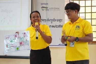 A teacher holds a microphone and a printed board game while a colleague stands beside her