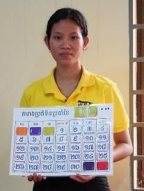 A teacher in a yellow shirt holds a large Khmer calendar board with colour-coded squares