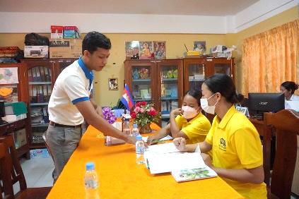 A man in a white shirt bends over a table to look at a book held by a woman