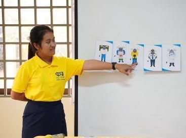 A teacher in a yellow shirt points to a row of occupation picture cards on a wall