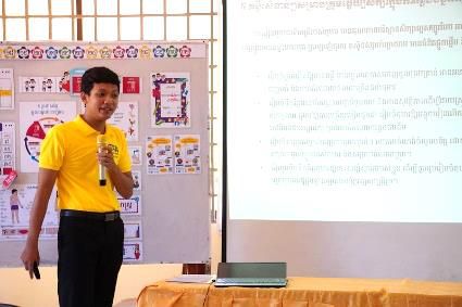 A man in a yellow shirt holds a microphone beside a display board and a projected slide
