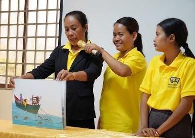 Three women stand behind a table as one holds a picture of a fishing boat for storytelling