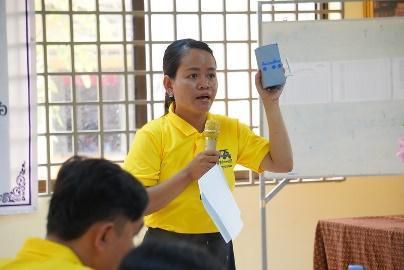 A trainer in a yellow shirt holds up a tablet while speaking into a microphone at the workshop