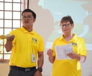 Two presenters in yellow shirts stand before a projected screen, one holding a microphone and papers