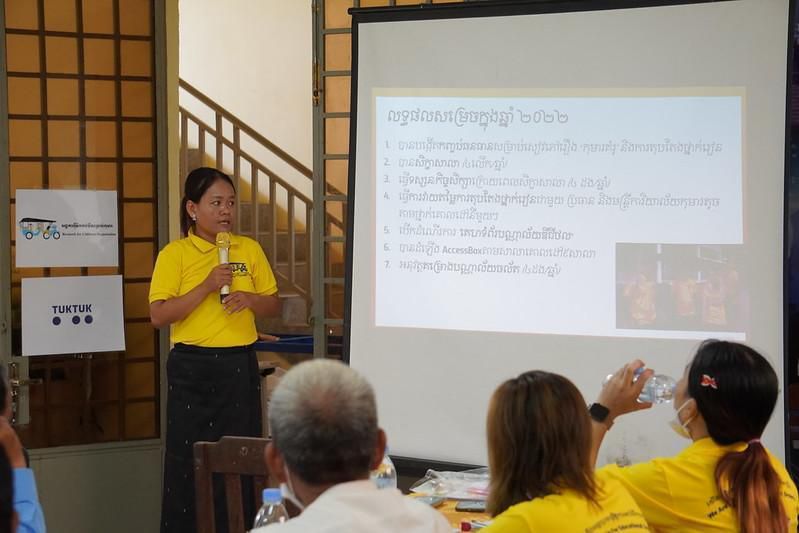 A woman in a yellow shirt presents the project results beside a projector screen at the annual meeting