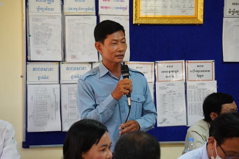 A man in a blue uniform shirt speaks into a microphone in front of a notice board at the meeting