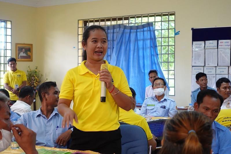 A woman in a yellow shirt speaks into a microphone to meeting participants seated around her