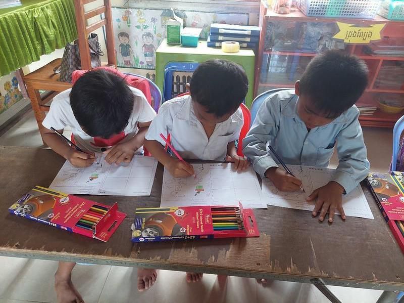 Three young students colour worksheets with colour pencils at a classroom table