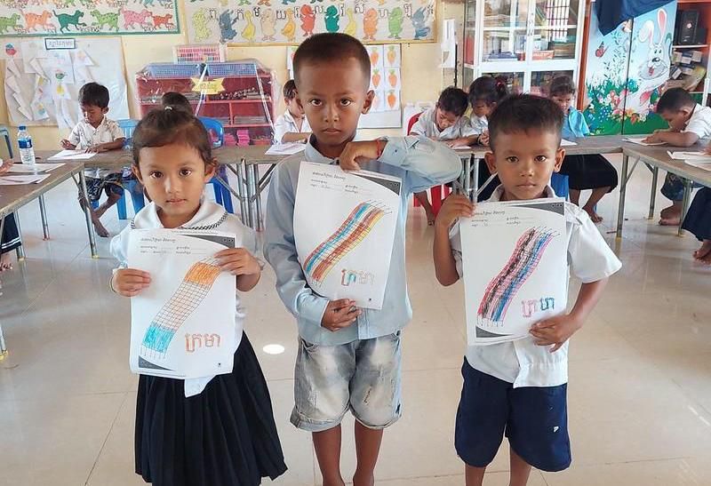 Three young students hold up colouring worksheets in their classroom