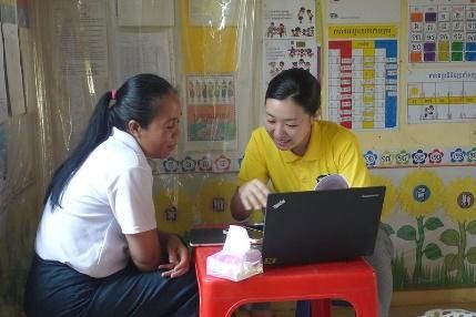 A staff member in a yellow shirt uses a laptop with a teacher during a classroom visit