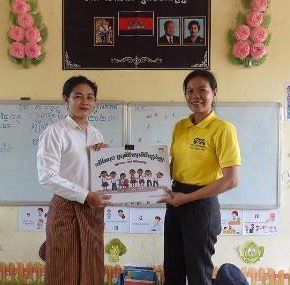 A teacher in a white shirt and a staff member in yellow hold up a poster in a classroom