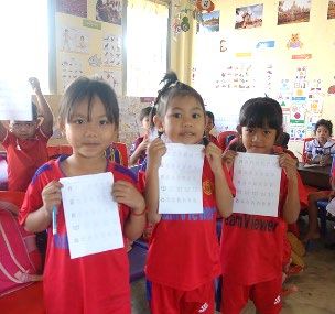 Three young students in red uniforms hold up worksheets they completed in their classroom