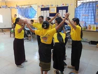 Workshop participants in yellow shirts dance in a circle with their arms raised during a song activity