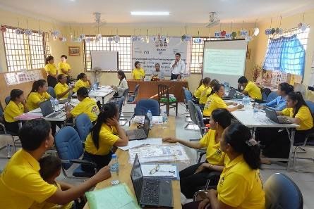 Teachers in yellow shirts sit at laptop tables in a large room during the fifth workshop