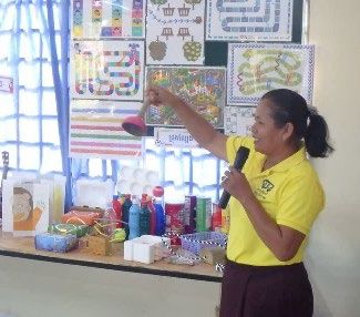 A trainer in a yellow shirt holds a microphone and points to classroom decoration materials displayed at the front