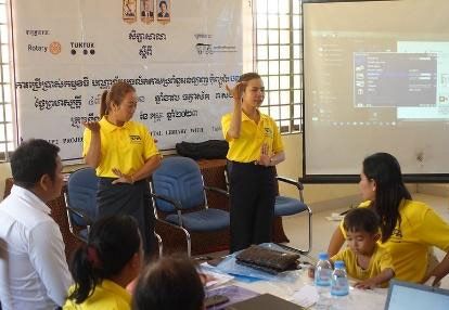 Participants in yellow shirts sit at tables facing a screen as two trainers stand to present