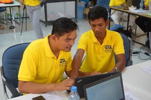 Two male teachers in yellow shirts work together on a laptop at a workshop table
