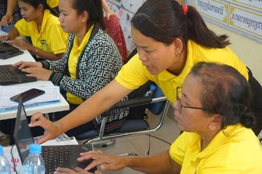 A trainer guides two teachers using laptops at a crowded workshop table