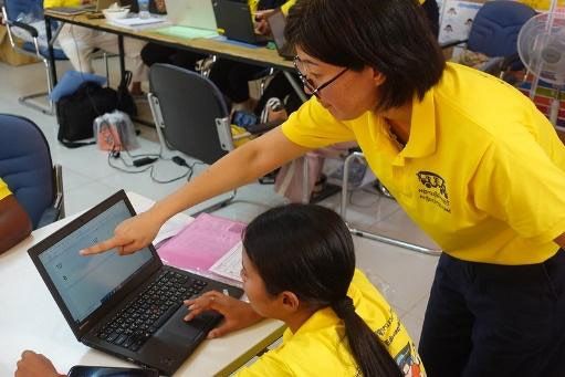 A trainer leans over to point at a laptop screen while a teacher practises making a worksheet