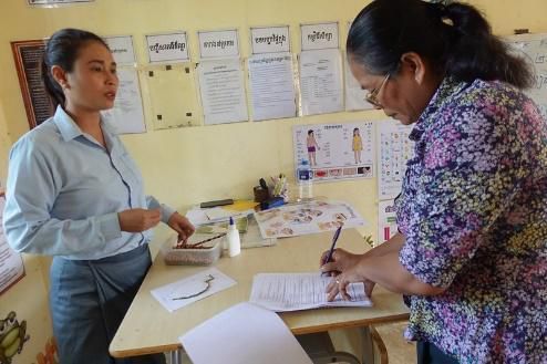 Two women stand at a classroom desk while one fills in an assessment sheet