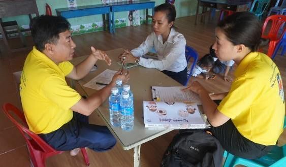 Two staff in yellow shirts sit at a small table with a teacher during a classroom decoration assessment