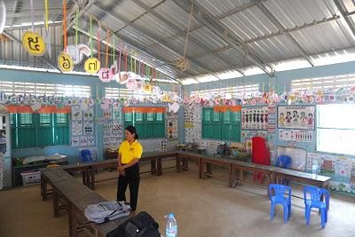 A decorated preschool classroom with hanging paper shapes, wall posters and low tables, with a teacher standing at the back