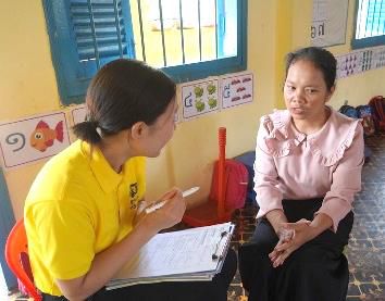 A staff member in yellow holds a clipboard and interviews a teacher in a classroom