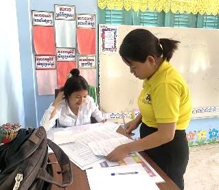 A staff member in yellow stands beside a seated teacher who looks over worksheets in a classroom
