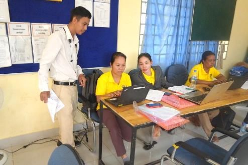 Teachers in yellow shirts practise at laptops around a table while a man in a white shirt stands beside them