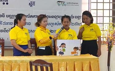 Four women in yellow shirts stand at a table while one speaks into a microphone holding a picture card