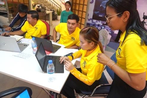 Teachers in yellow shirts work on laptops around a white table during the sixth workshop