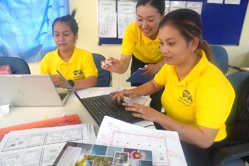 A trainer leans over to guide two teachers who are making a worksheet on a laptop
