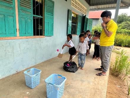 Young children toss balls into baskets in a school corridor as a man in yellow watches