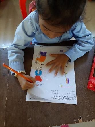 A young child colours in a printed worksheet with an orange pencil at a wooden desk