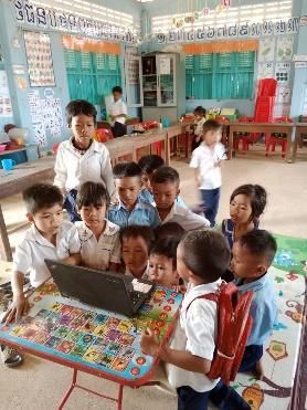 Several young children crowd around a laptop placed on a colourful table in a classroom