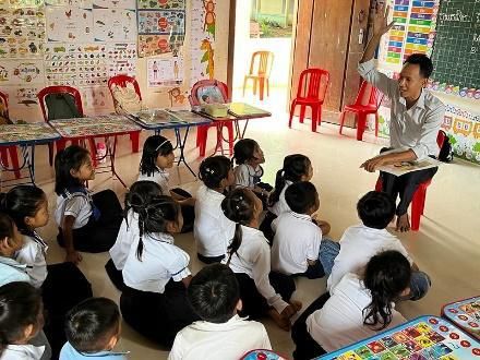 Young pupils in school uniform sit on a classroom floor while a teacher leads an activity