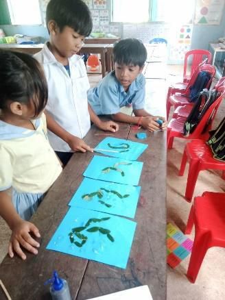 Young children press green leaves onto blue paper sheets on a long wooden table during a craft activity