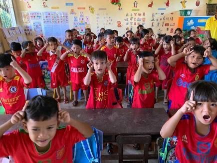 Preschool children in red shirts raise their hands to their ears while singing in a crowded classroom